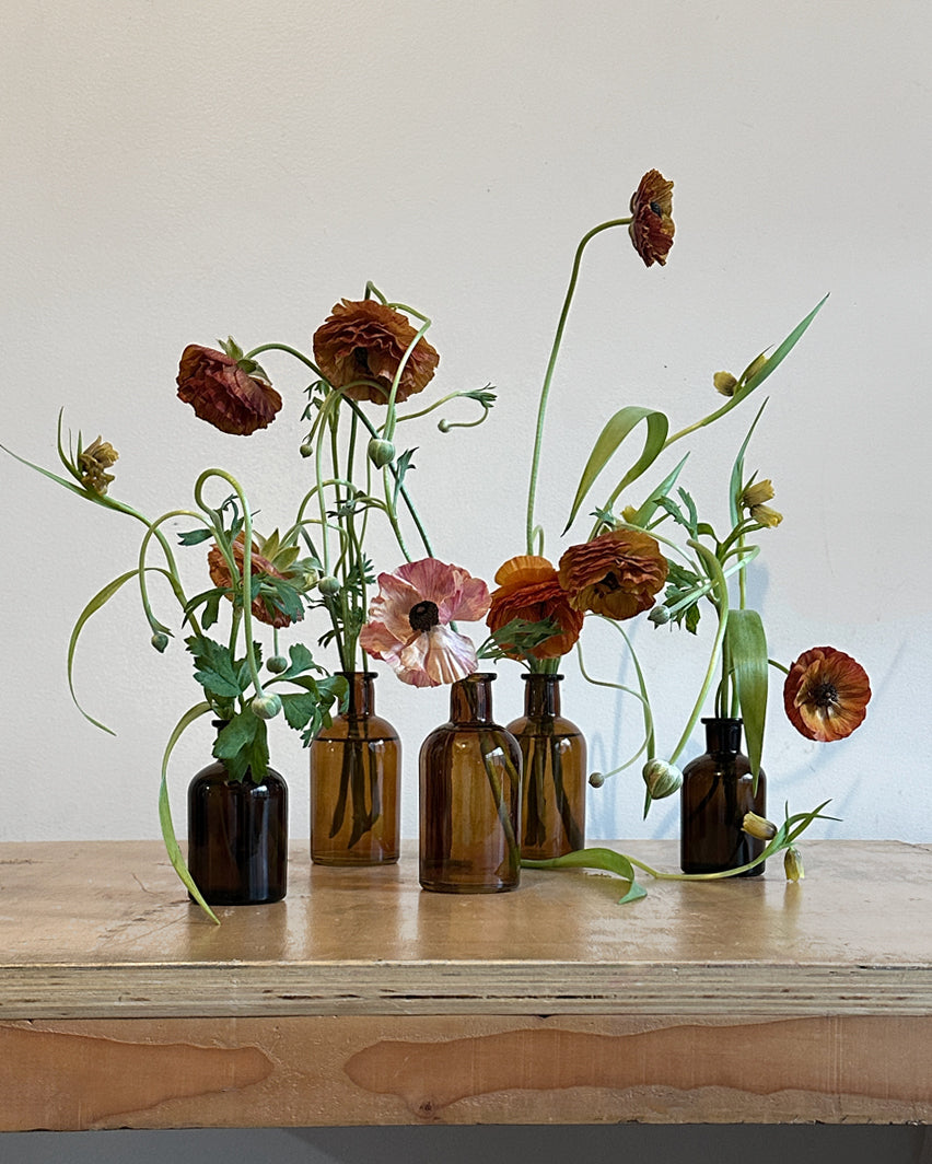 Set of five amber glass bud vases, each containing an assortment of seasonal flowers.