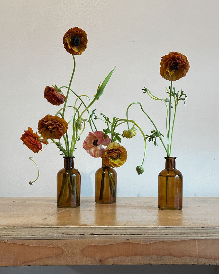 Set of three amber glass bud vases, each containing an assortment of seasonal flowers.